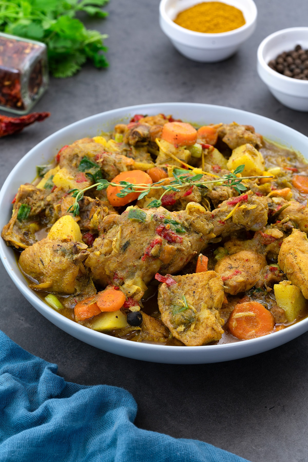 Jamaican curry chicken served in a white bowl on a grey table. Accompanied by cups of turmeric powder and allspice berries, as well as a small green and blue towel.