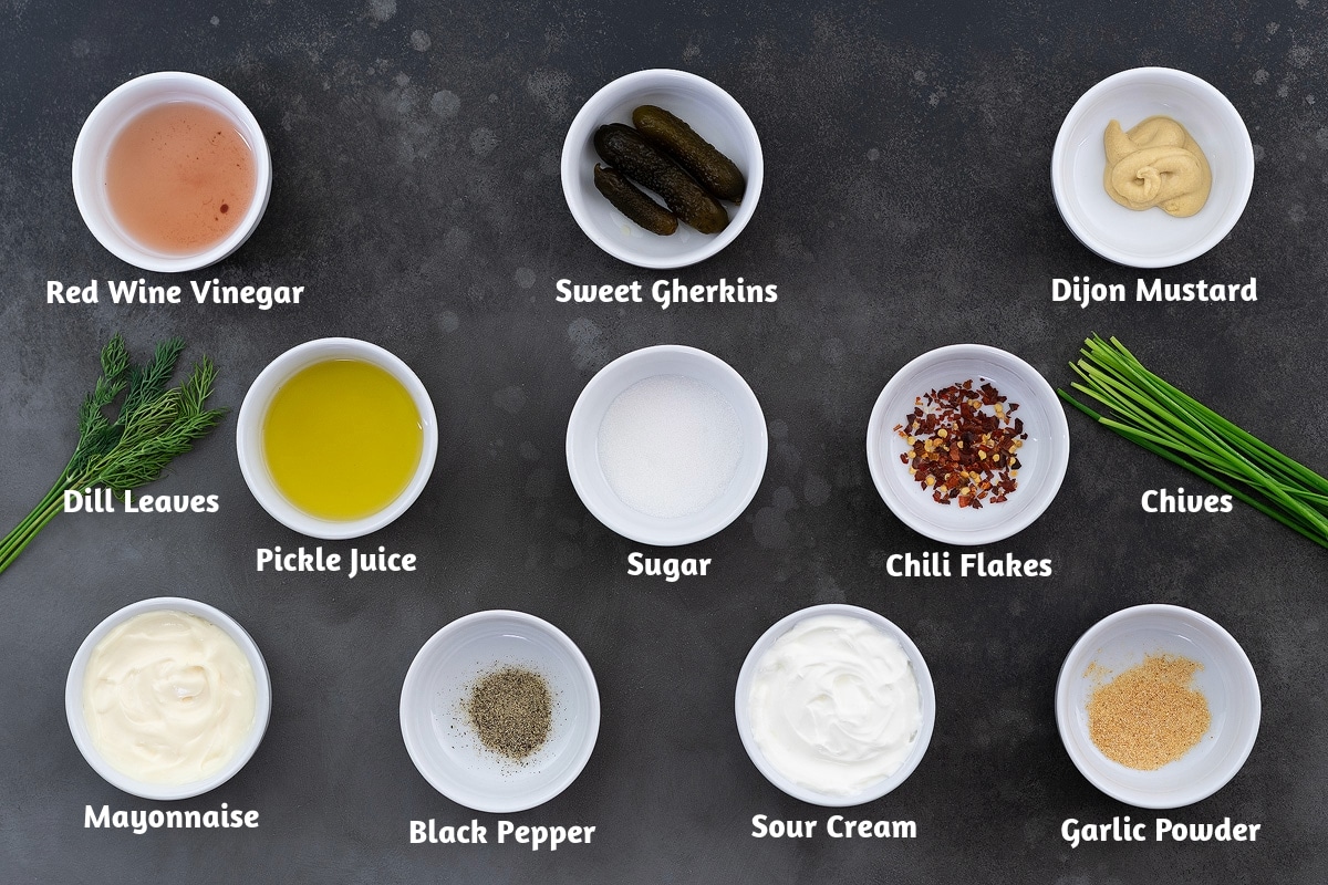 Macaroni salad dressing ingredients creatively arranged on a grey table: red wine vinegar, sweet gherkins, Dijon mustard, dill leaves, pickle juice, sugar, chili flakes, chives, mayonnaise, black pepper, sour cream, and garlic powder.