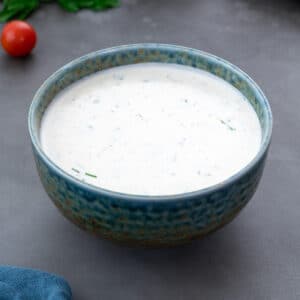 Appetizing fresh homemade ranch dressing in a green and blue patterned bowl on a grey table. Parsley leaves and cherry tomatoes are arranged around the bowl.