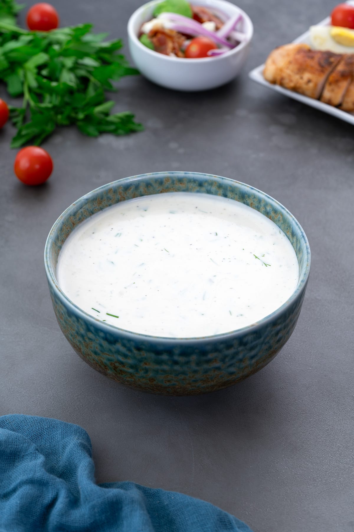 Appetizing fresh homemade ranch dressing in a green and blue patterned bowl on a grey table. Parsley leaves and cherry tomatoes are arranged around the bowl, accompanied by salad and chicken breast in a small cup and a plate.
