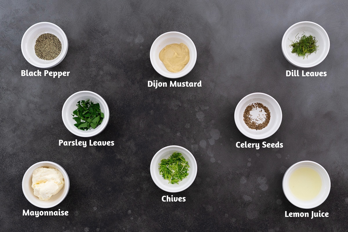 Arrangement of dressing ingredients on a grey table: In the first row, black pepper, Dijon mustard, and dill leaves; in the second row, parsley leaves and celery seeds; and in the third row, mayonnaise, chives, and lemon juice.