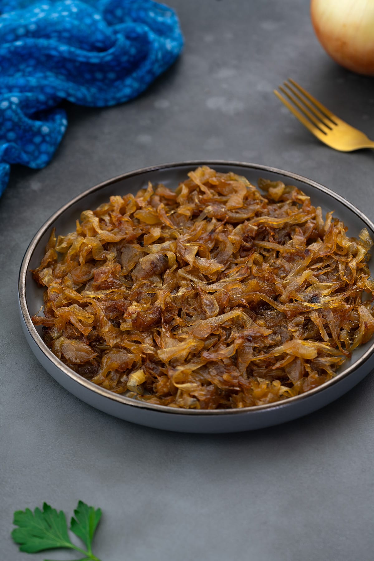 Perfect Caramelized Onions served on a light gray plate, placed on a gray table and surrounded by a golden spoon, onions, and a blue towel.