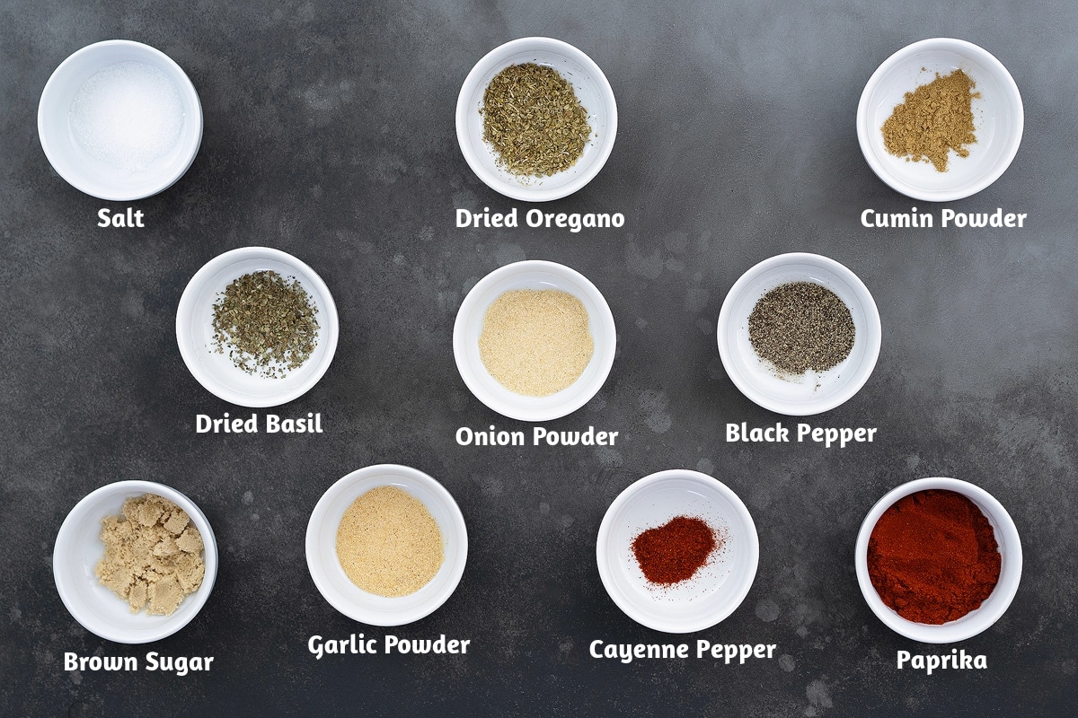 Chicken seasoning mix or dry rub recipe ingredients on a gray table: salt, dried oregano, cumin powder, dried basil, onion powder, black pepper powder, brown sugar, garlic powder, cayenne pepper, and paprika.