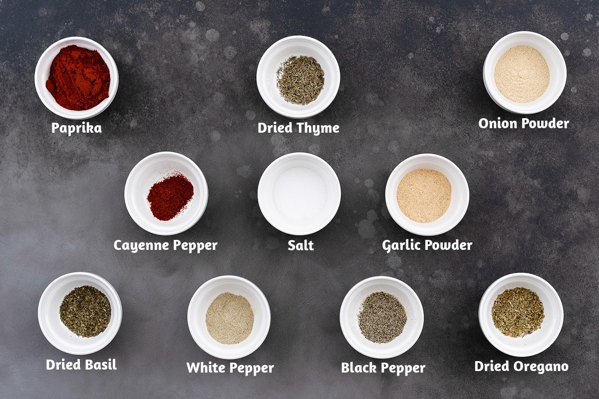 White cups containing the ingredients for Creole seasoning: paprika, dried thyme, onion powder, cayenne pepper, salt, garlic powder, dried basil, white pepper powder, black pepper powder, and dried oregano. The cups are arranged on a gray table.