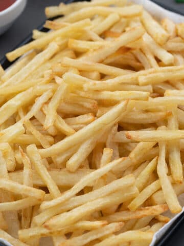 Golden Crispy Homemade French Fries in a black tray on a grey table with a cup of ketchup placed nearby.