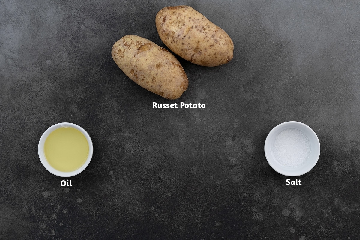 Ingredients for French fries recipe, including russet potatoes, oil, and salt, arranged on a grey table.