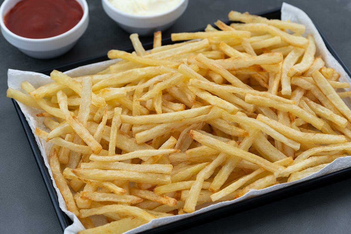 Golden Crispy Homemade French Fries in a black tray on a grey table with a cup of ketchup and mayonnaise placed nearby.