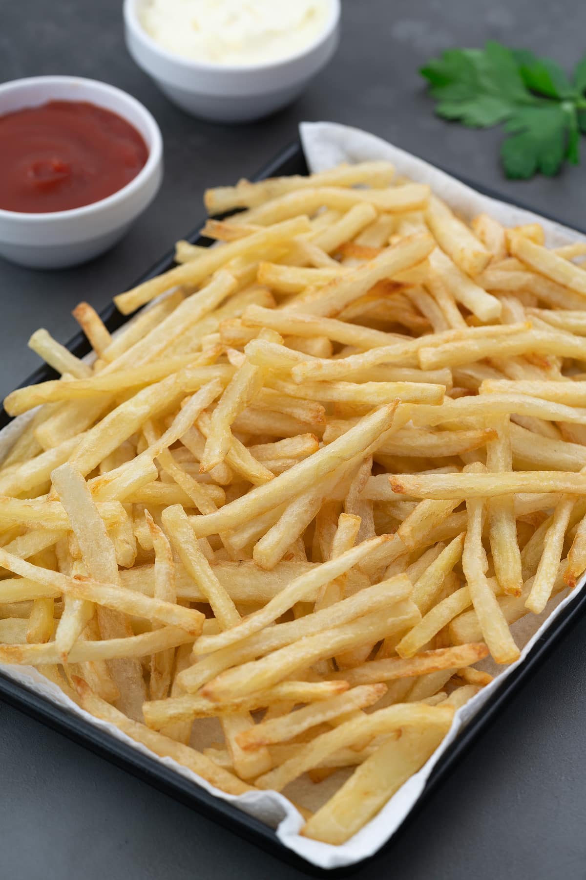 Golden Crispy Homemade French Fries in a black tray on a grey table with a cup of ketchup and mayonnaise placed nearby.