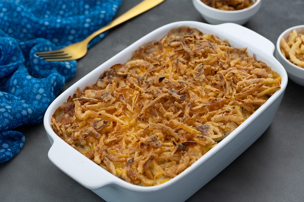 Homemade Green Bean Casserole in a white baking dish on a grey table, accompanied by French-fried onions, caramelized onions in cups, a blue towel, and a golden spoon nearby.