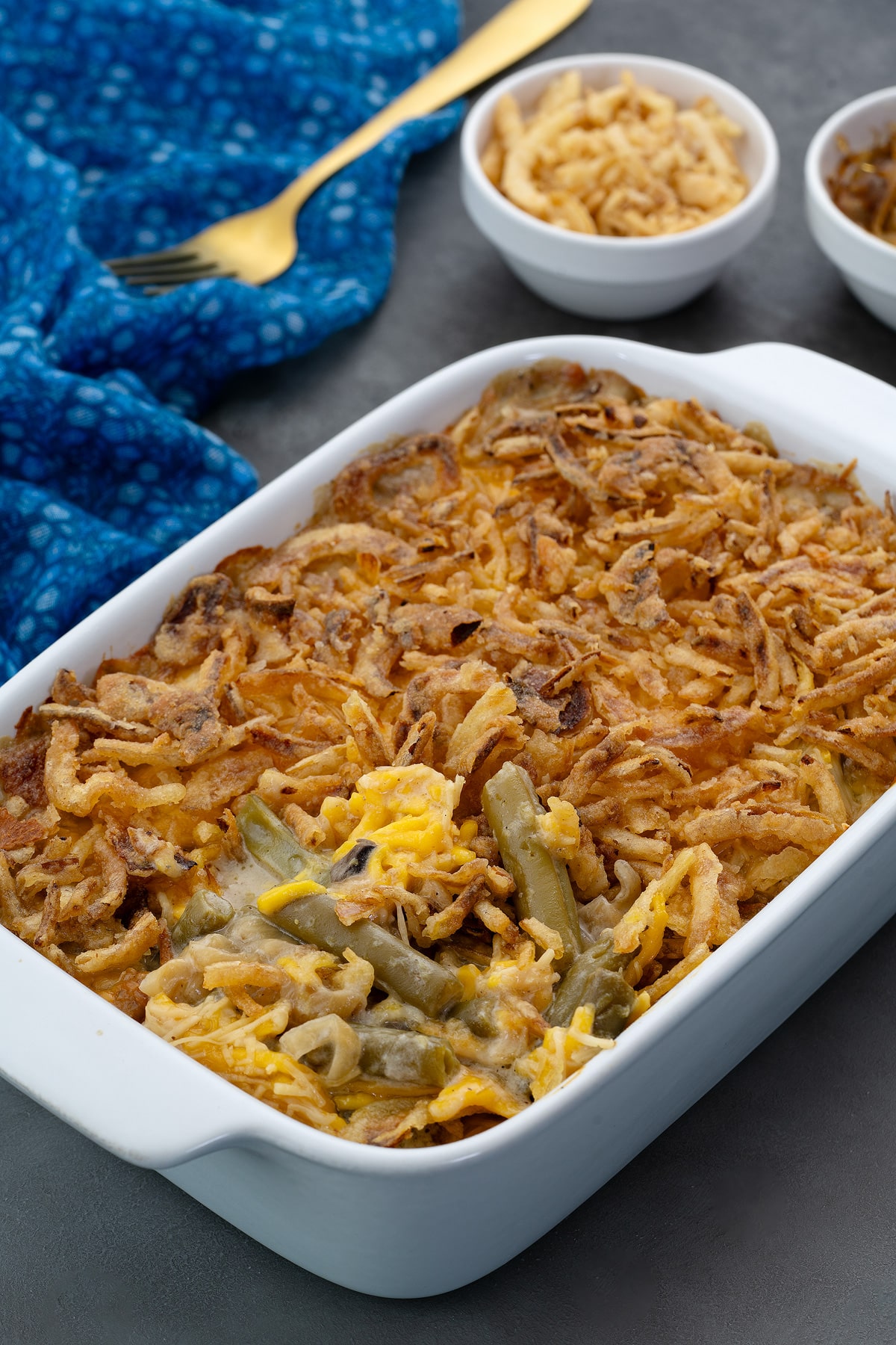 Homemade Green Bean Casserole in a white baking dish on a grey table, accompanied by French-fried onions, caramelized onions in cups, a blue towel, and a golden spoon nearby.