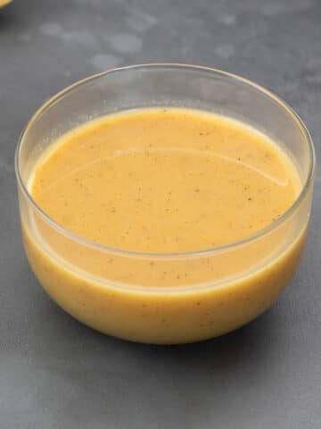 Honey mustard dressing in a glass bowl on a gray table with a cup of honey, lemon slice, and greens arranged around it.