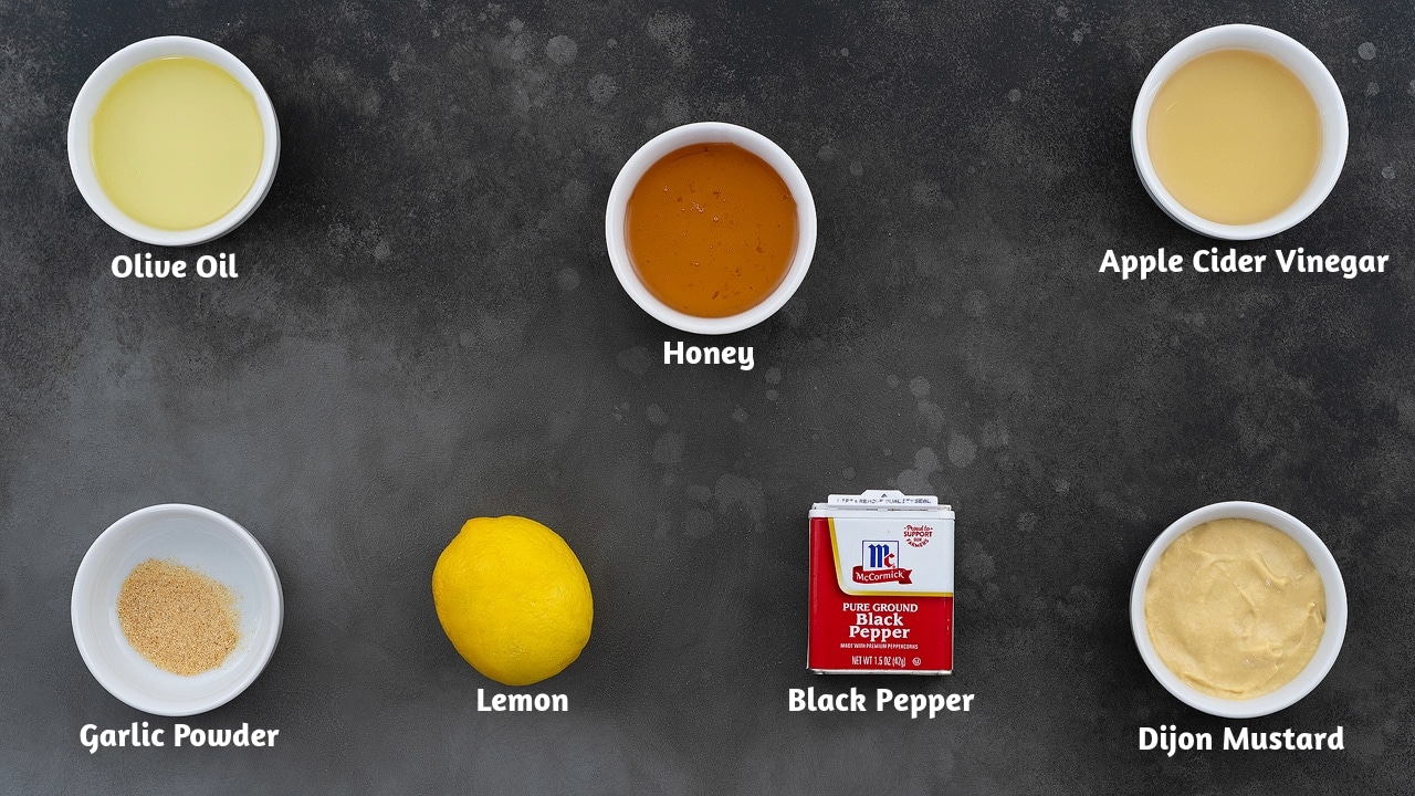 Honey mustard dressing ingredients are arranged in a row on a gray table. It consists of Olive Oil, Honey, and Apple Cider Vinegar, Garlic Powder, Lemon, Black Pepper, and Dijon Mustard.