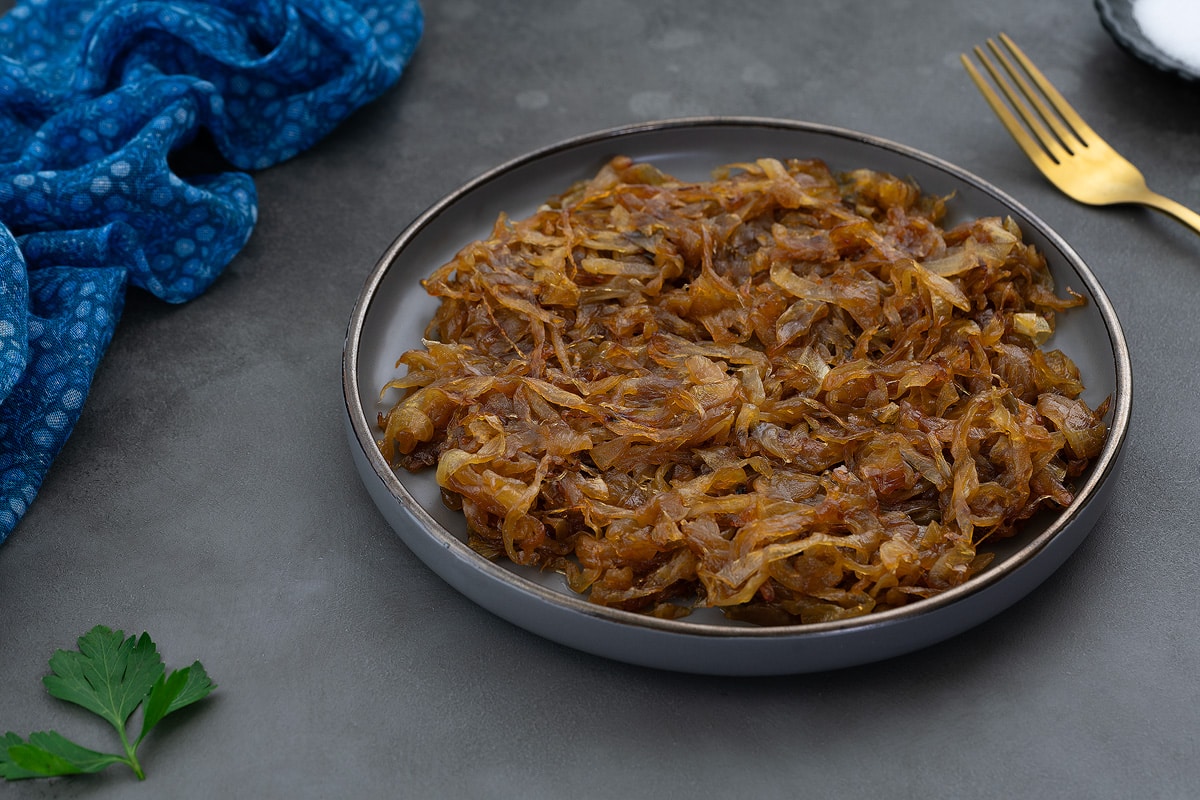 Perfect Caramelized Onions served on a light gray plate, placed on a gray table and surrounded by a golden spoon, and a blue towel.