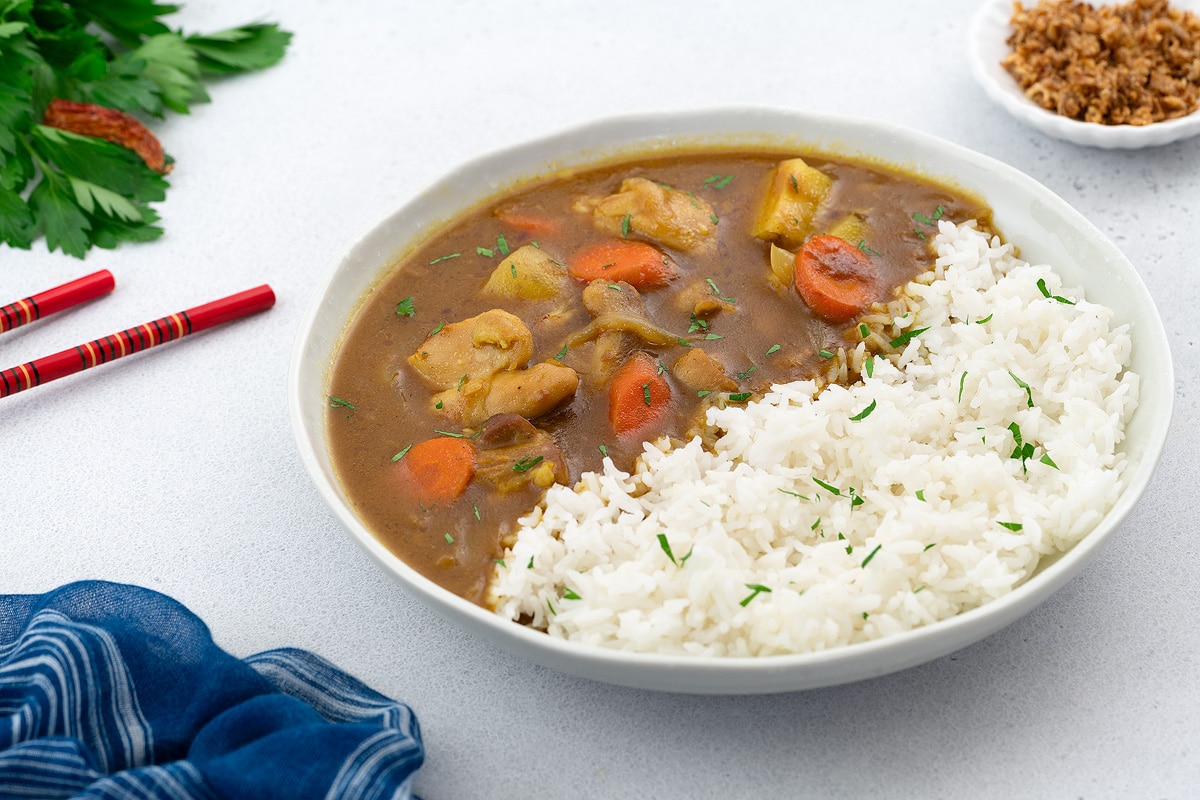 Japanese chicken curry served in a white ceramic bowl with a side of rice, presented on a white tabletop. Accompanied by chopsticks, onions, garlic cloves, and assorted greens arranged around.