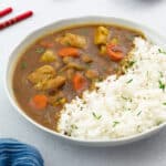 Japanese chicken curry served in a white ceramic bowl with a side of rice, presented on a white tabletop.