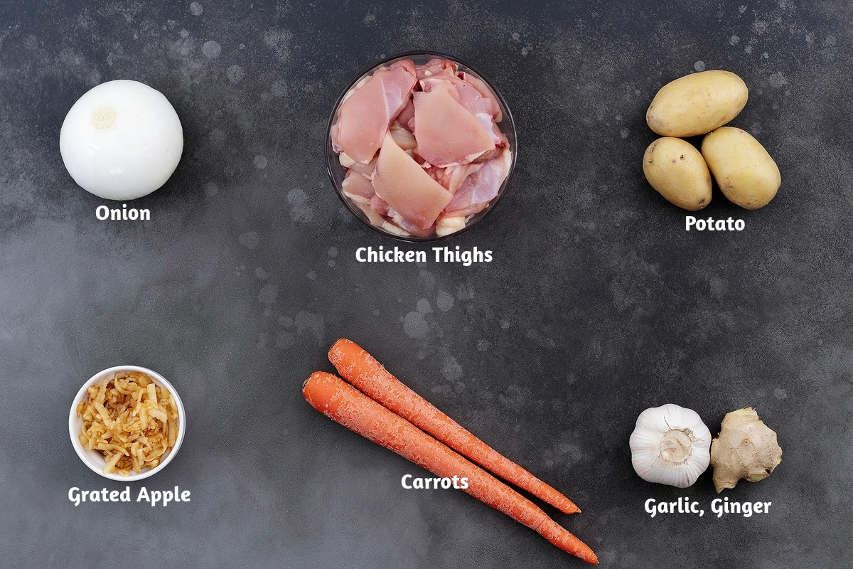 An assortment of Japanese curry ingredients on a grey tabletop, including onions, chicken thighs, potatoes, grated apples, carrots, ginger, and garlic.