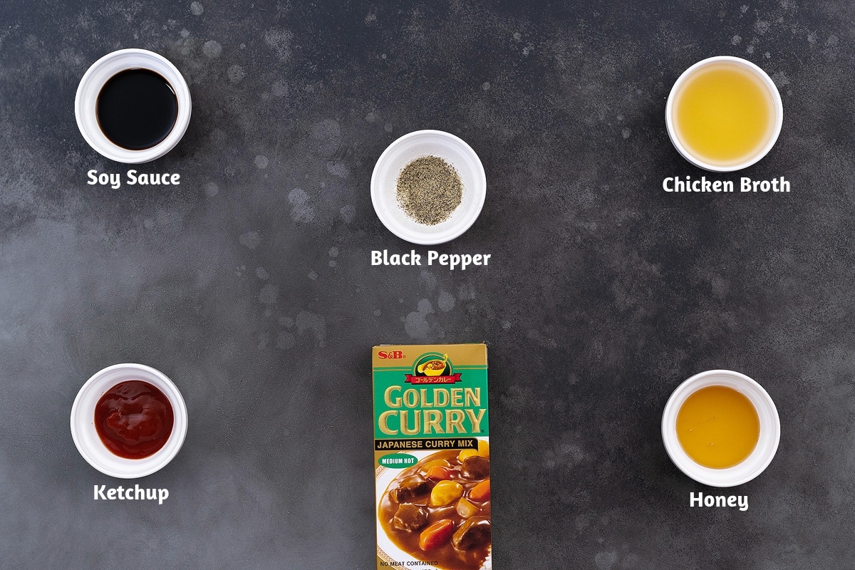 A selection of Japanese curry seasonings and flavorings arranged on a grey table, featuring soy sauce, black pepper, chicken broth, ketchup, Japanese curry roux, and honey.