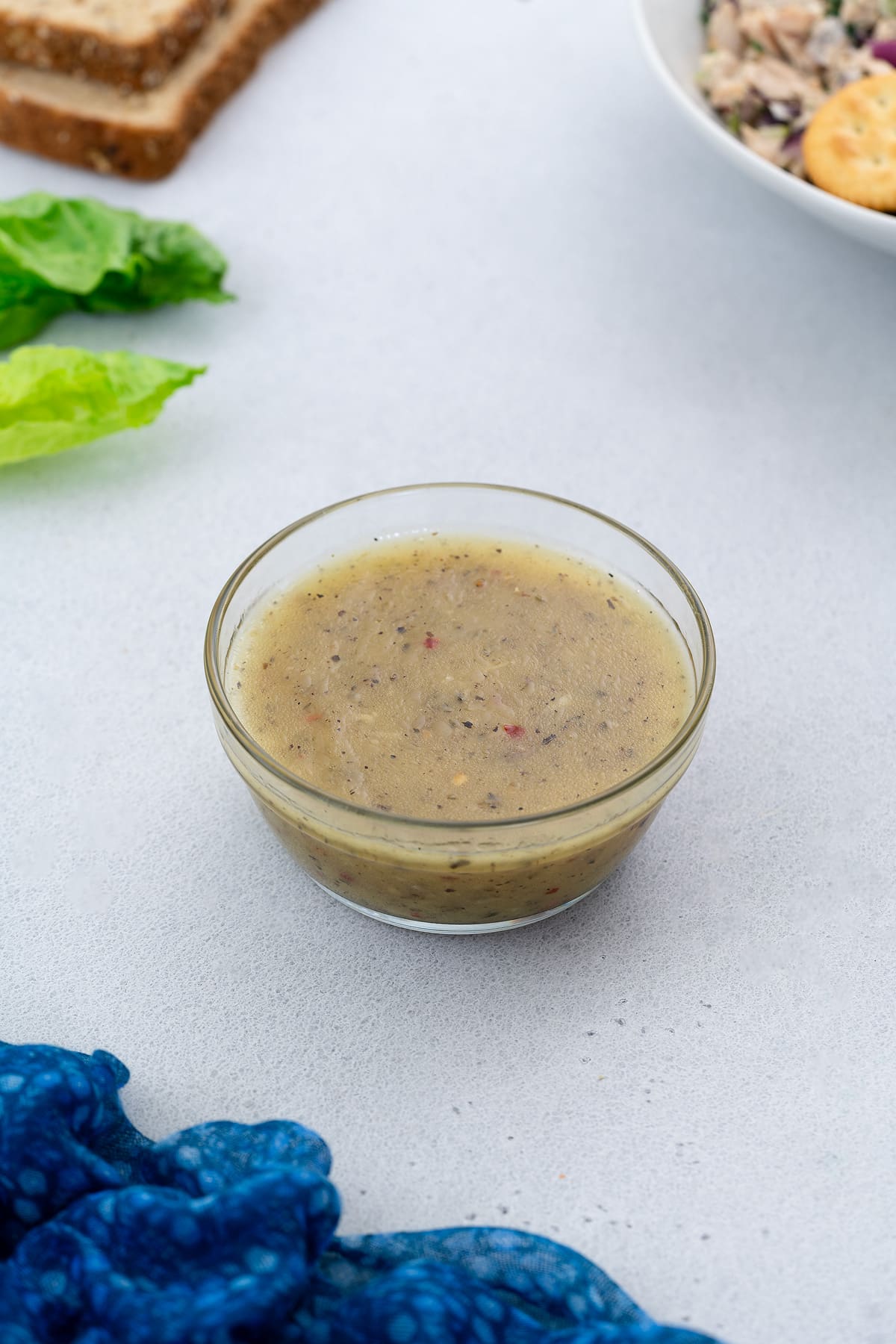 Salad dressing in a glass bowl on a white table with salad, bread slices, and lettuce leaves arranged around it.