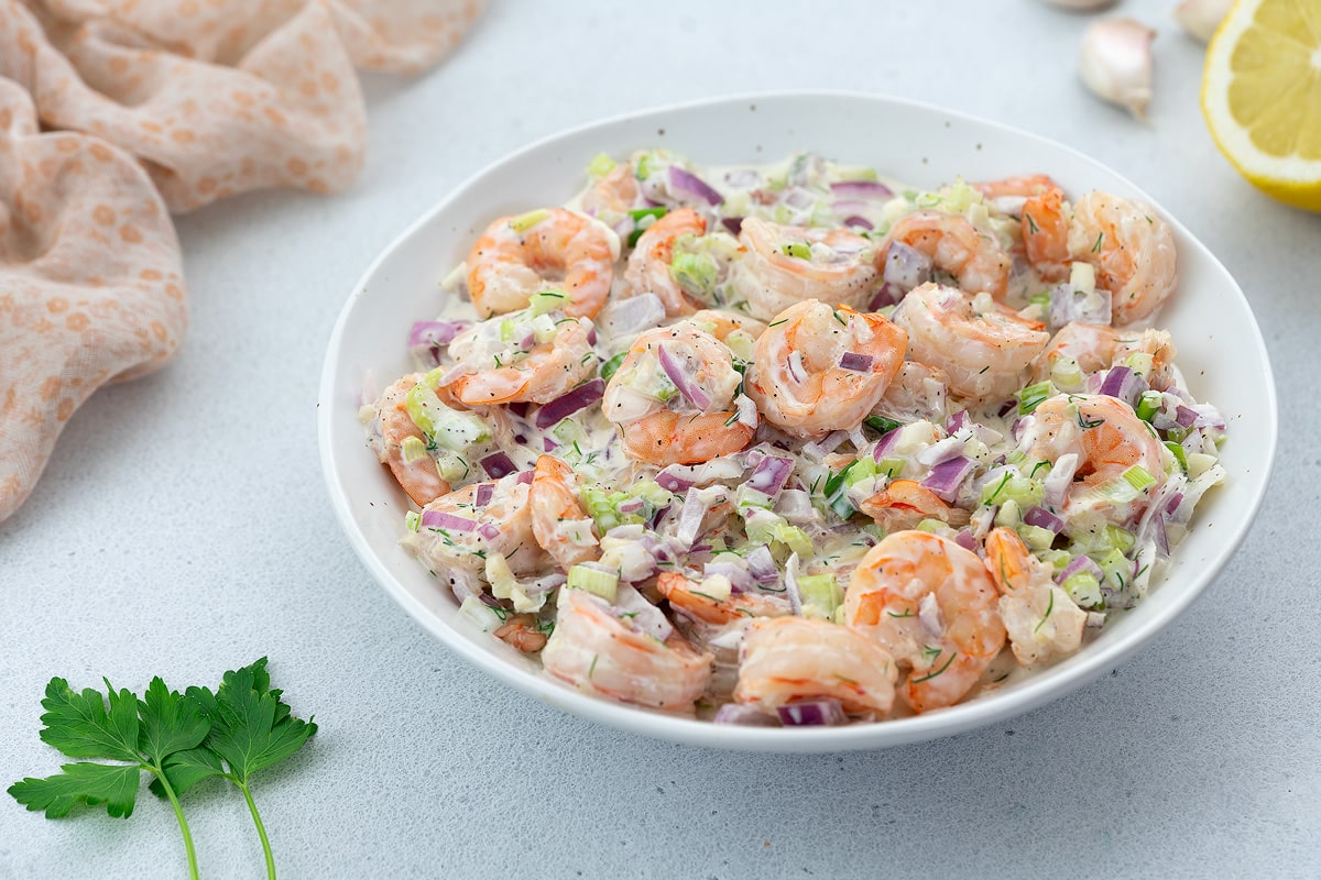 Creamy homemade shrimp salad in a white bowl on a white tabletop, surrounded by garlic cloves and a towel.