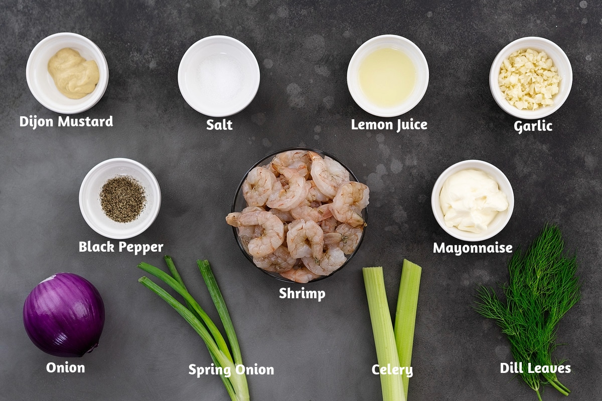Ingredients for shrimp salad arranged on a grey table. The ingredients include Dijon mustard, salt, lemon juice, garlic, black pepper powder, shrimp, mayonnaise, onion, spring onion, celery, and dill leaves.