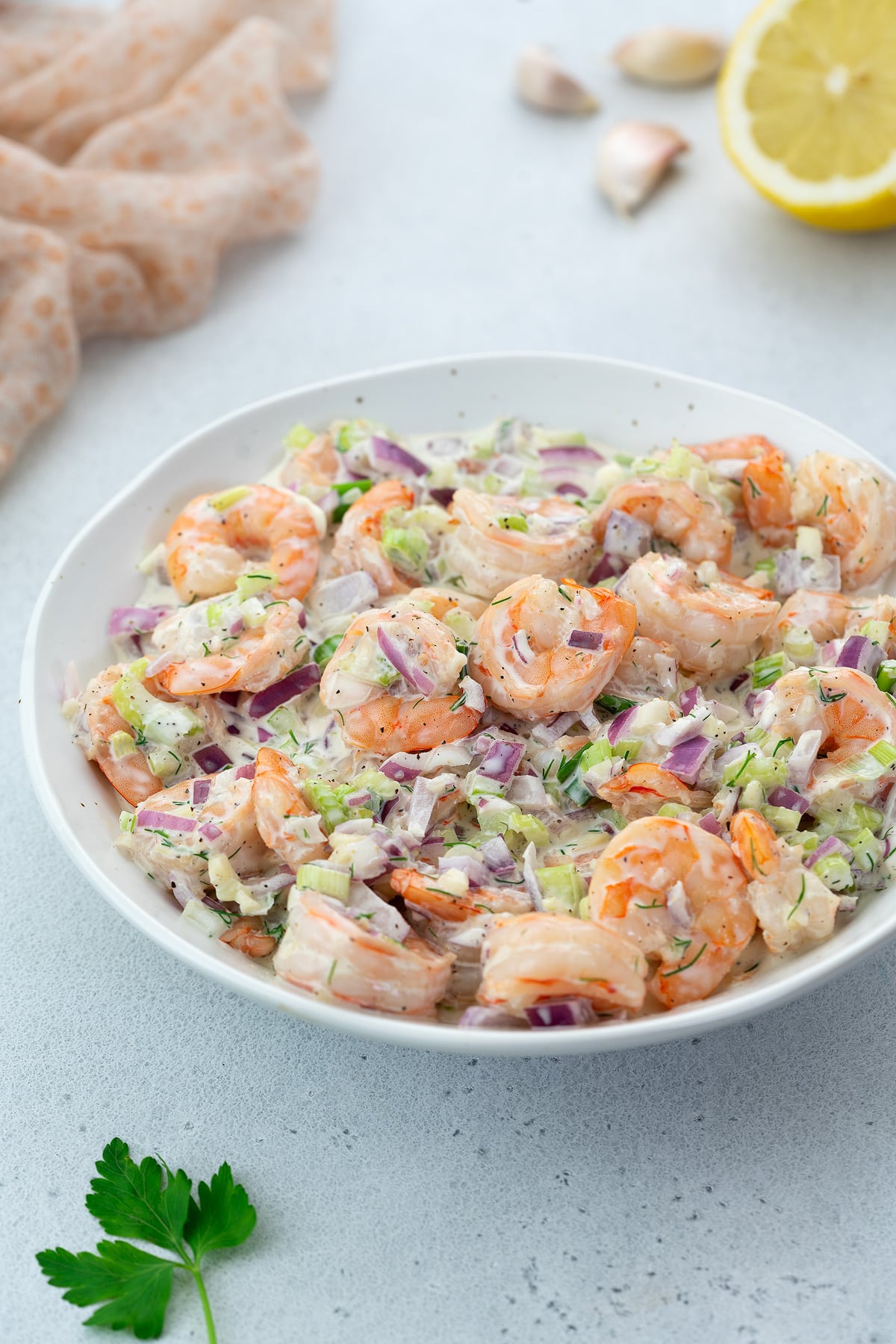 Creamy homemade shrimp salad in a white bowl on a white tabletop, surrounded by garlic cloves and a towel.
