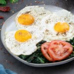 Sunny-side up eggs on a white plate, with kale and tomato slices.