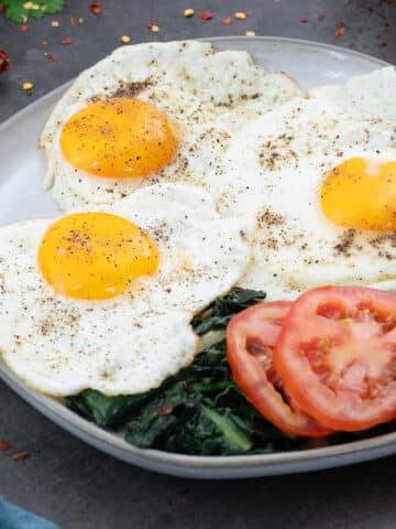 Sunny-side up eggs on a white plate, with kale and tomato slices.