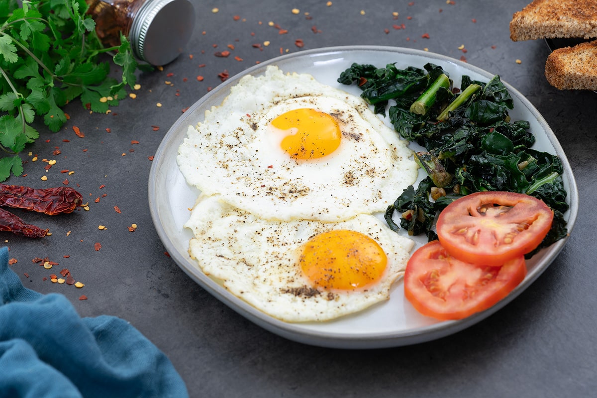 Sunny-side up eggs on a white plate, with kale and tomato slices, bread, and greens placed around.