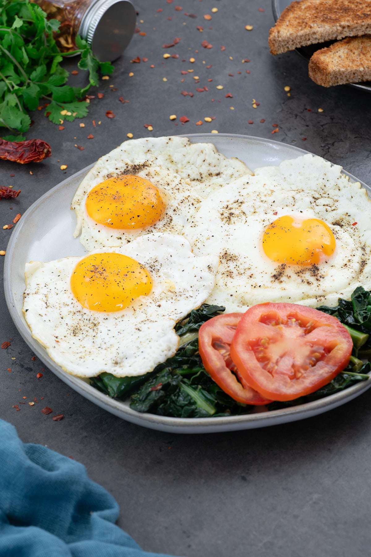 Sunny-side up eggs on a white plate, with kale and tomato slices, bread, and greens placed around.