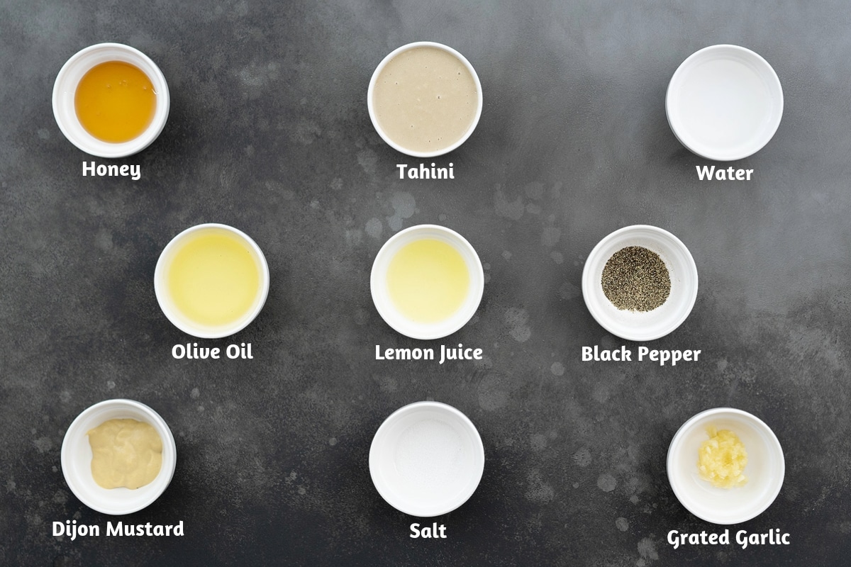 A gray table with the ingredients for tahini dressing: honey, tahini, water, olive oil, lemon juice, black pepper powder, Dijon mustard, salt, and grated garlic.