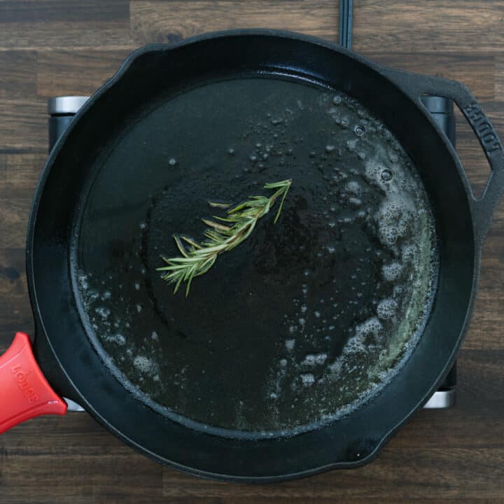 An iron skillet with rosemary infusing in oil butter mixture.