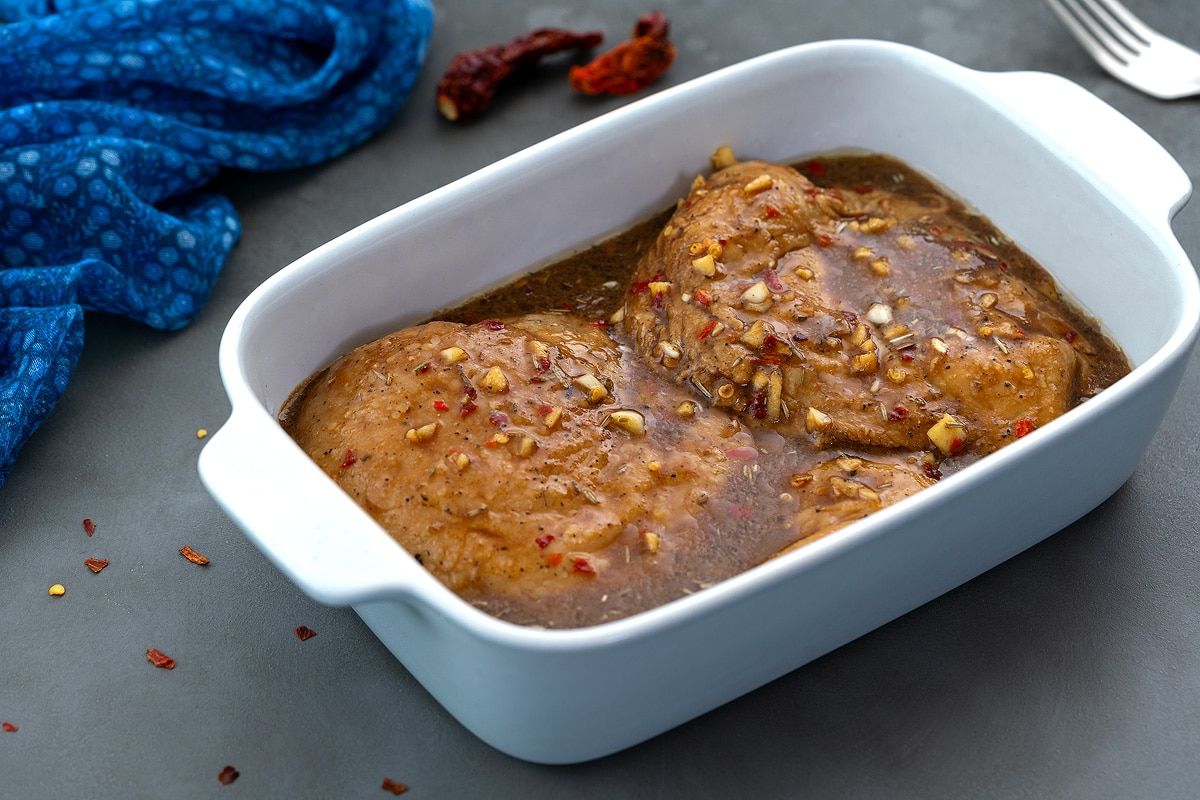 Chicken marinade in a white dish, mixed with chicken breasts, placed on a grey table, with a blue towel and a fork nearby.