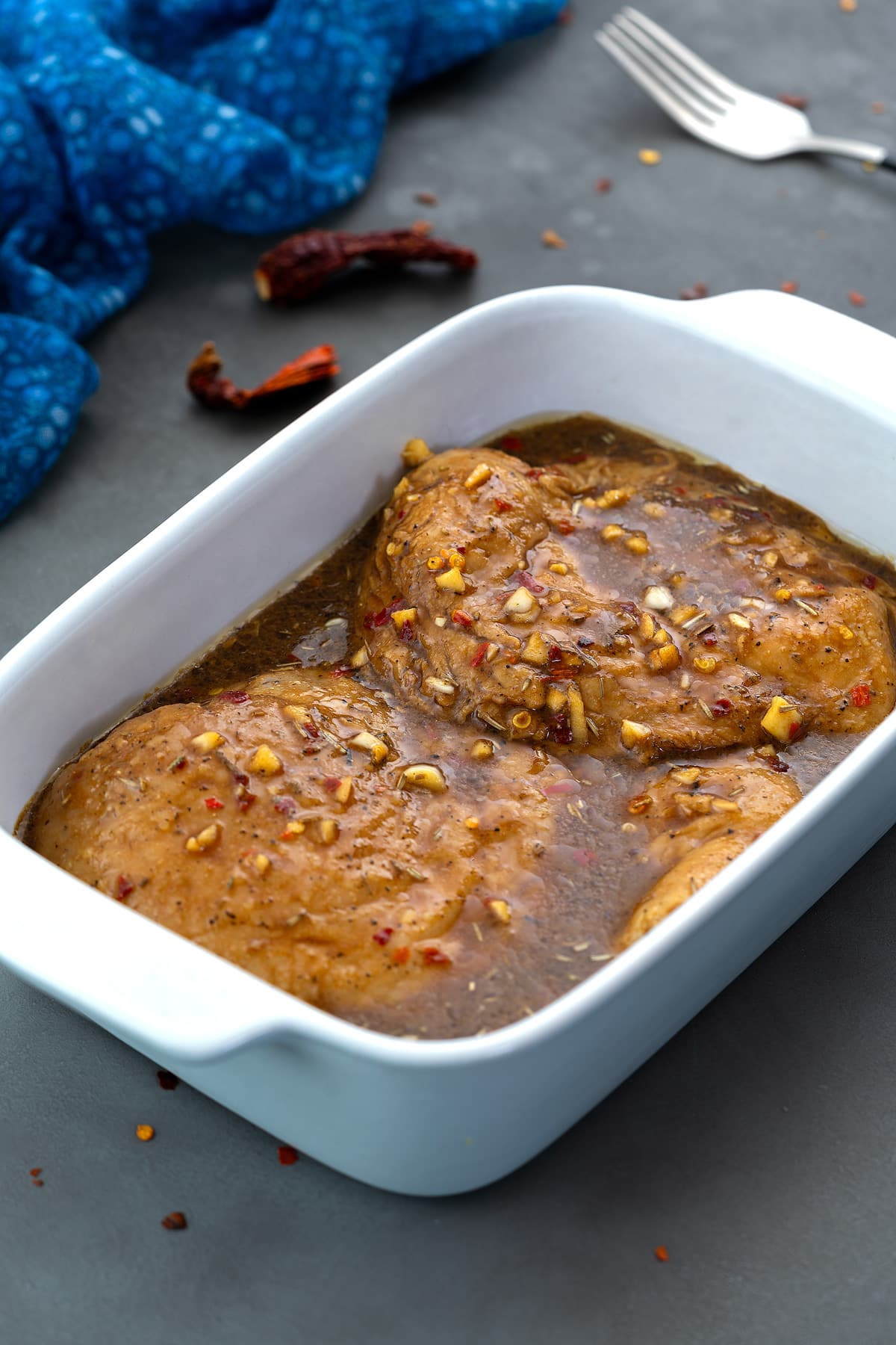 Chicken marinade in a white dish, mixed with chicken breasts, placed on a grey table, with a blue towel and a fork nearby.