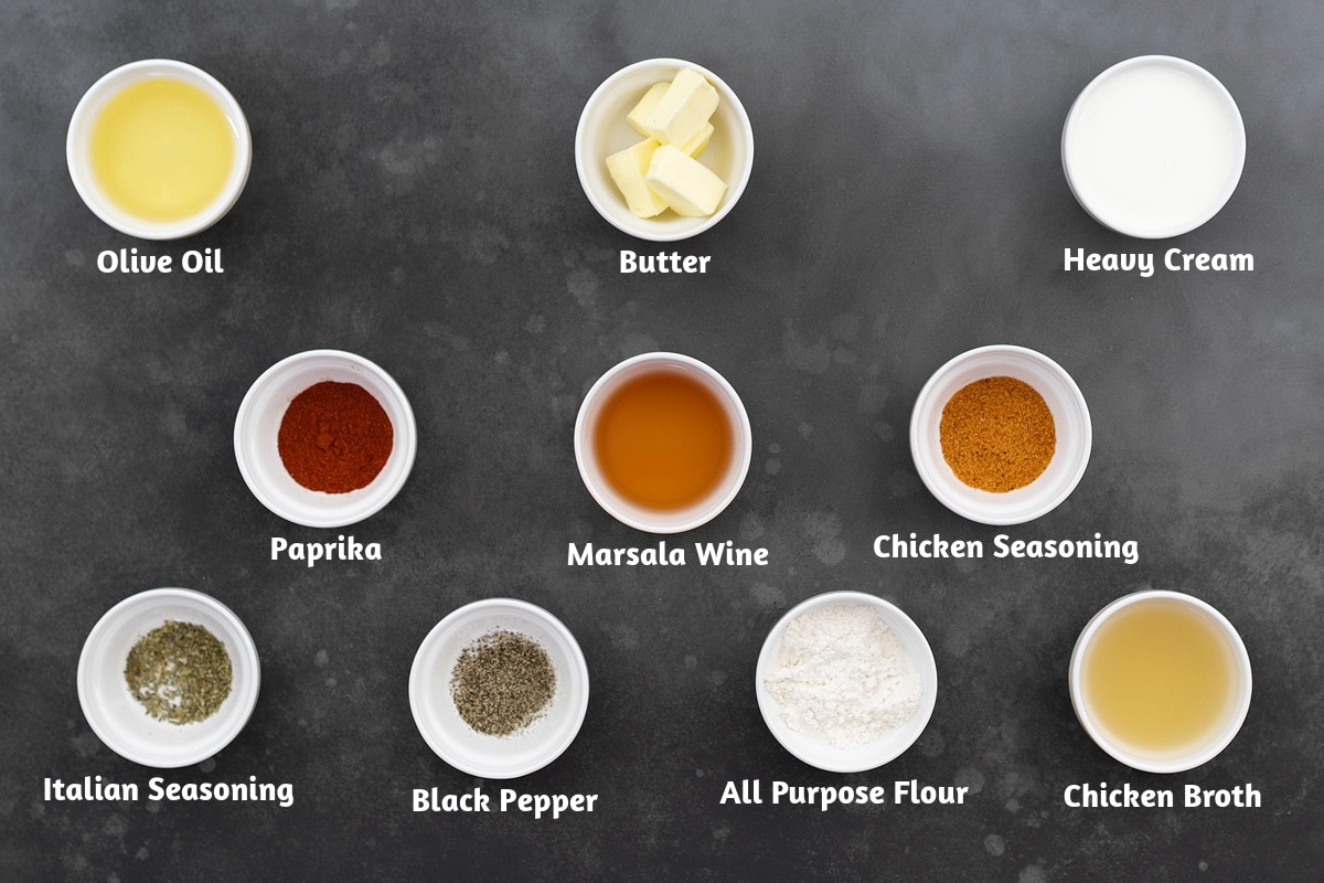 A grey table with ingredients for chicken marsala, including olive oil, butter, heavy cream, paprika, Marsala wine, chicken seasoning, Italian seasoning, black pepper, all-purpose flour, and chicken broth.