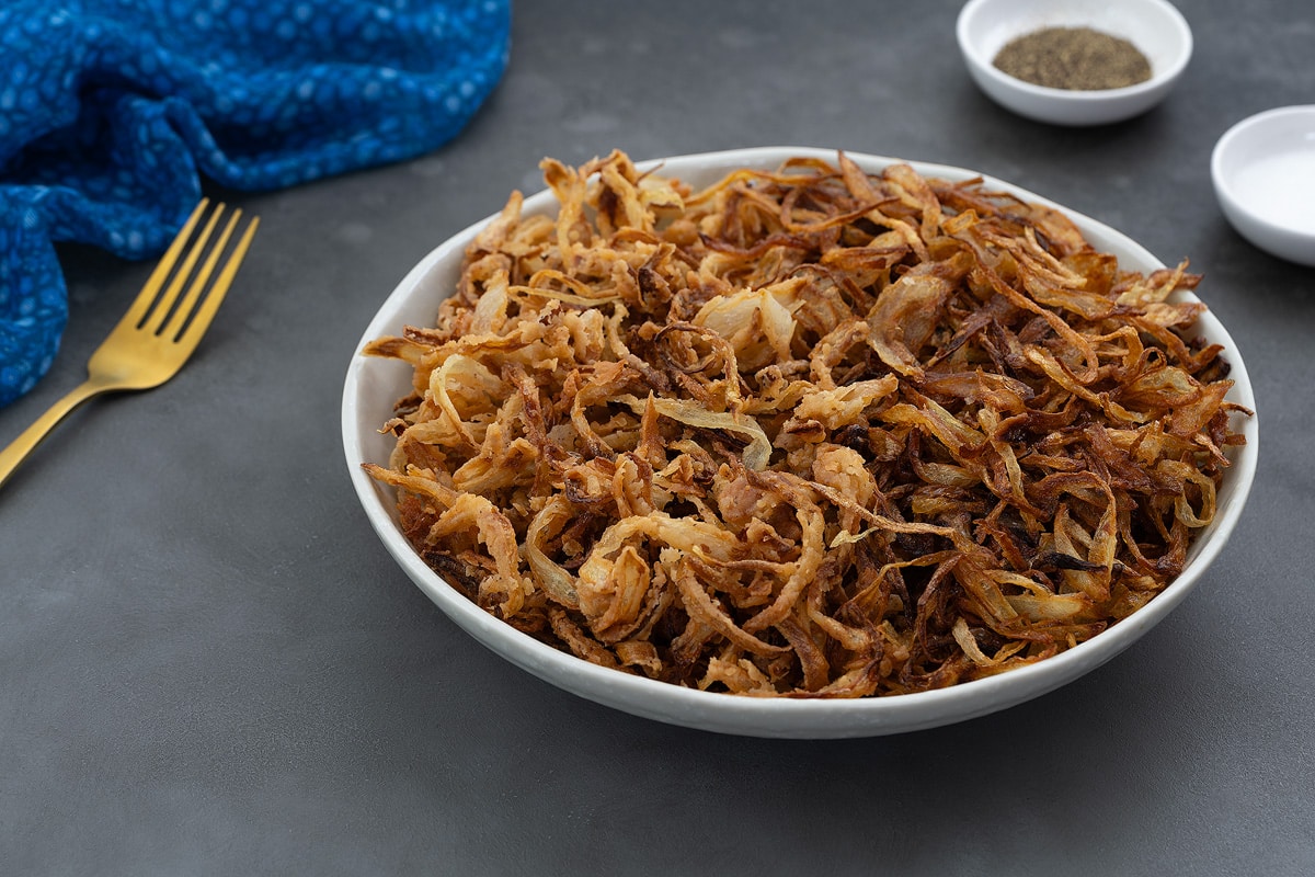 Classic and French fried onions in a white bowl placed on a gray table. A blue towel, golden fork, and salt and pepper powder in small white plates placed around.