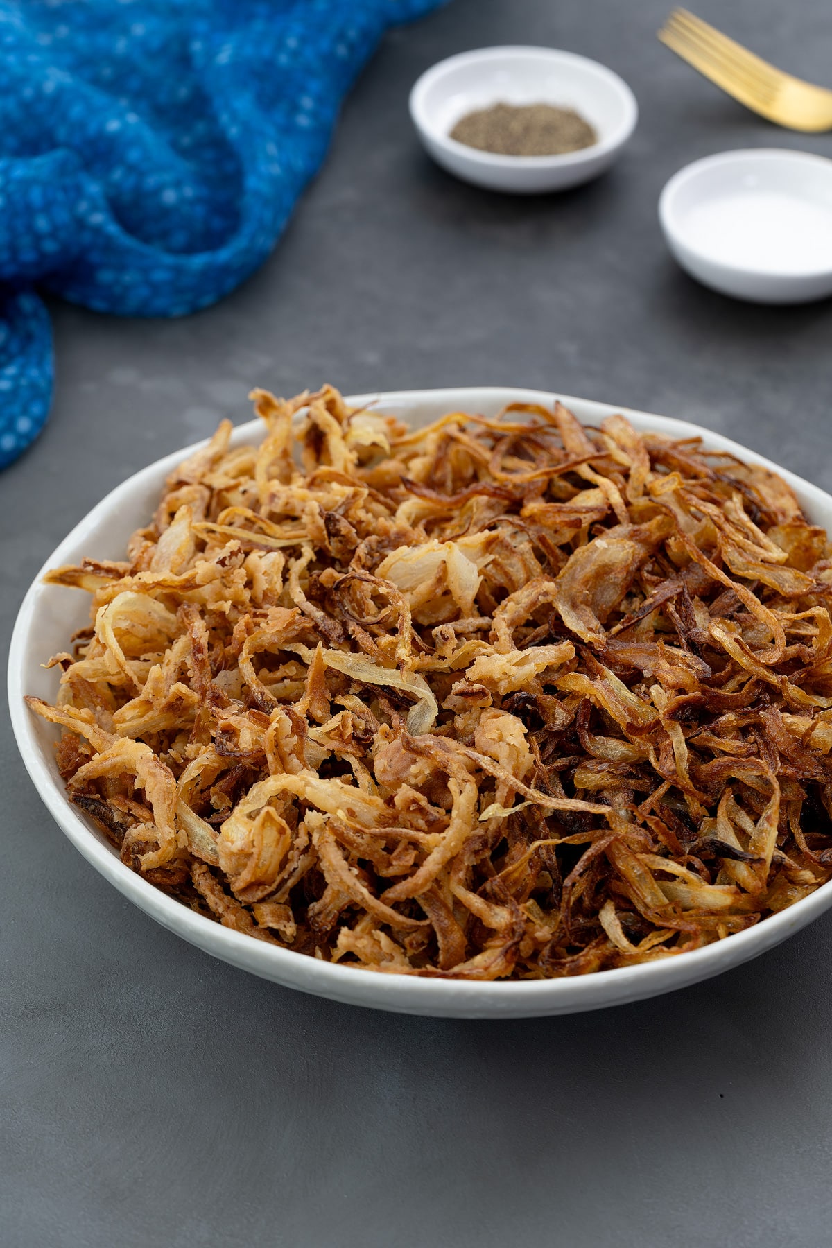 Classic and French fried onions in a white bowl placed on a gray table. A blue towel, golden fork, and salt and pepper powder in small white plates placed around.