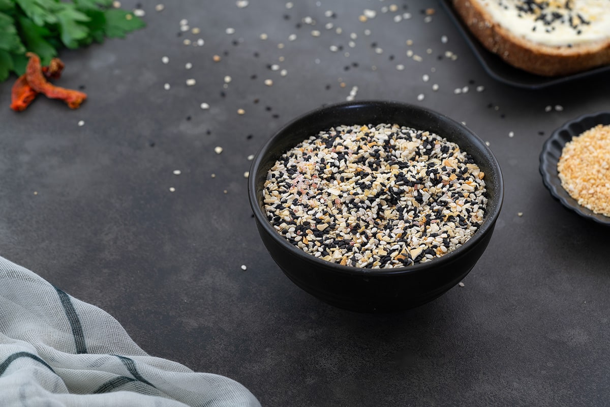 Everything bagel seasoning mix on a black plate placed on a grey table. Accompanied by dried minced garlic on a small plate, slices of bread, and parsley leaves arranged around it.