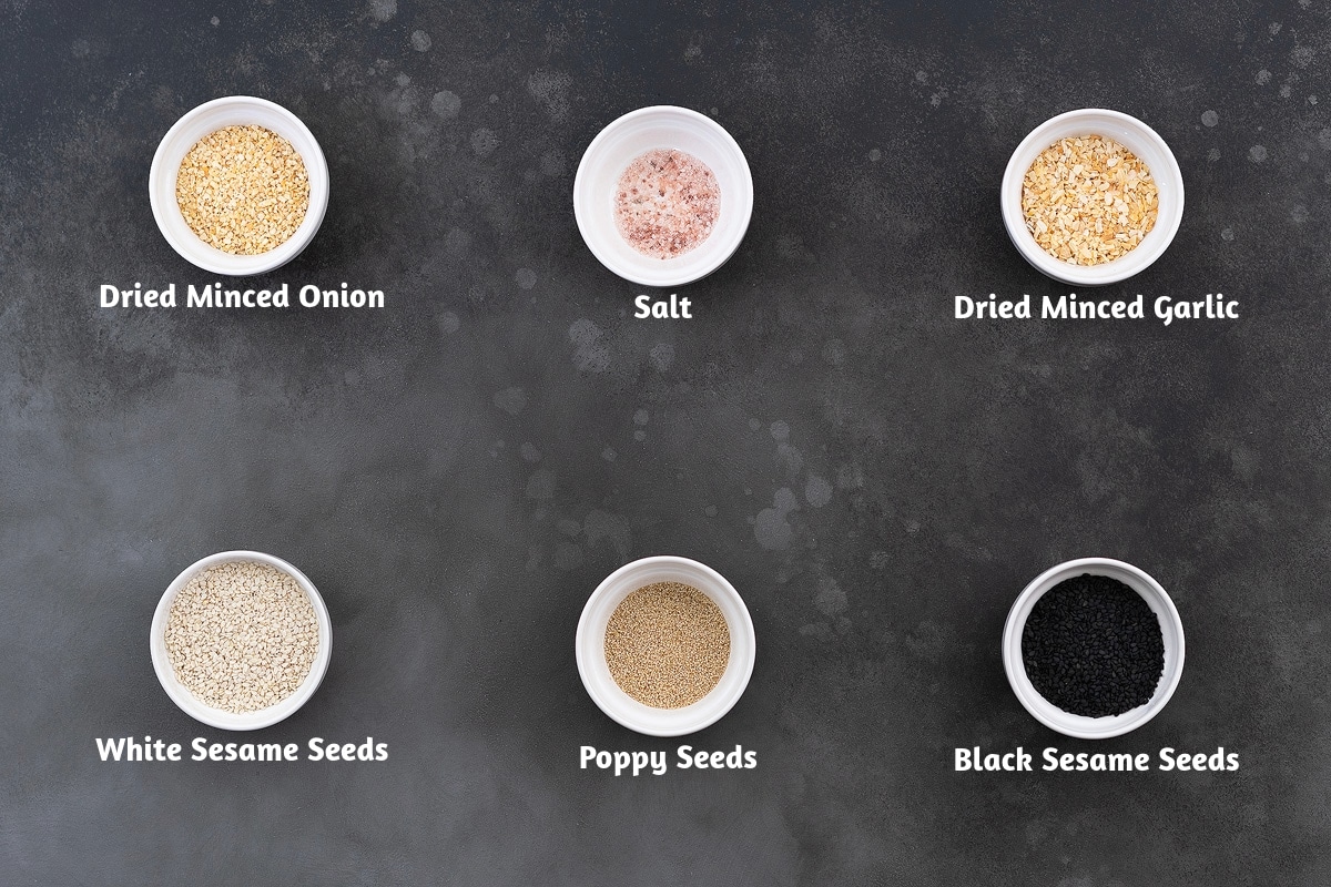 Everything bagel seasoning ingredients arranged on a grey table. The ingredients include dried minced onion, salt, dried minced garlic, white sesame seeds, poppy seeds, and black sesame seeds.