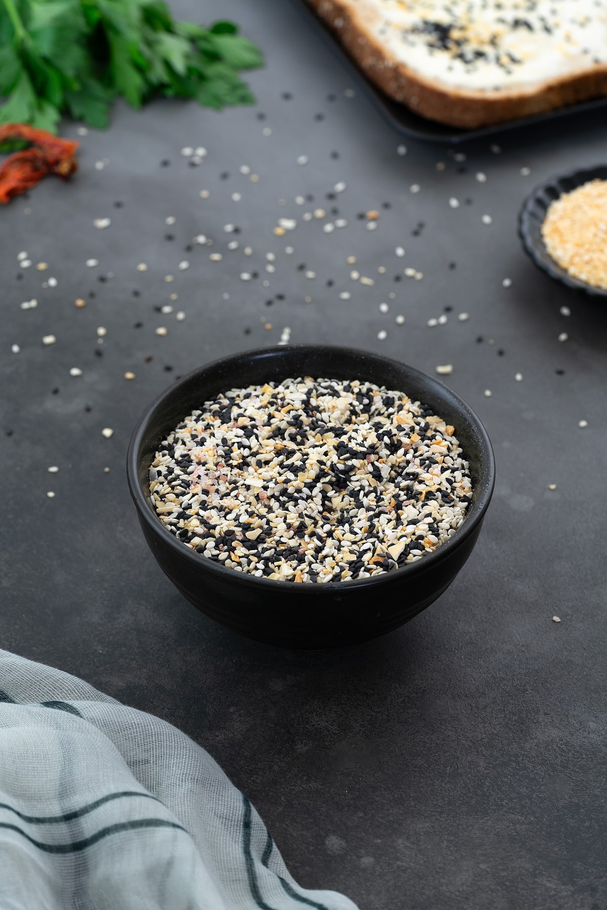 Everything bagel seasoning mix on a black plate placed on a grey table. Accompanied by dried minced garlic on a small plate, slices of bread, and parsley leaves arranged around it.