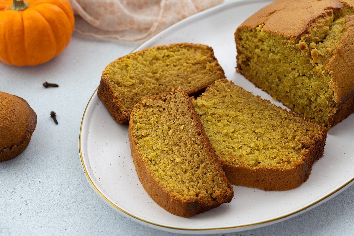 Three slices of pumpkin bread on an oval white plate on a white table, with a small pumpkin, pumpkin cup cake, and a golden fork placed nearby.