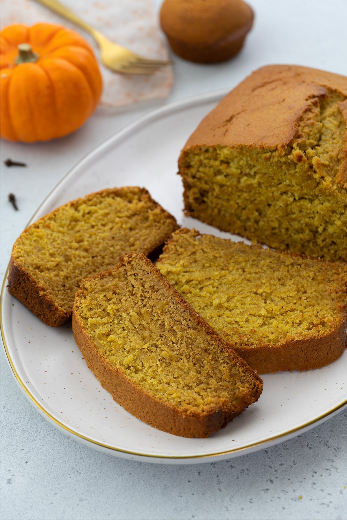 Three slices of pumpkin bread on an oval white plate on a white table, with a small pumpkin, pumpkin cup cake, and a golden fork placed nearby.