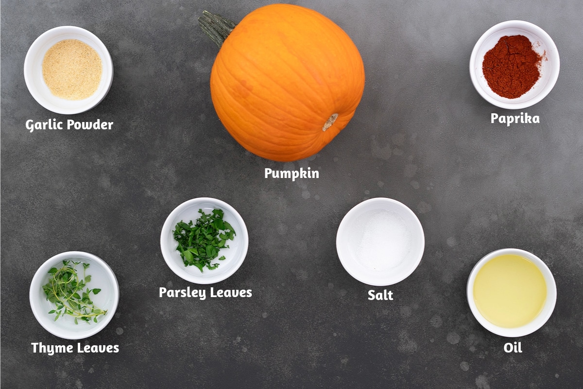 
Roasted pumpkin ingredients on a gray table: garlic powder, pumpkin, paprika, thyme leaves, parsley leaves, salt, and oil.