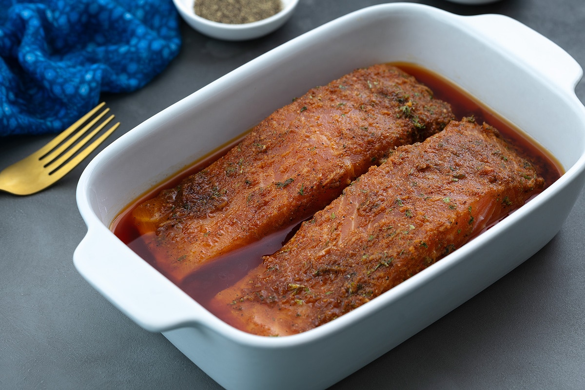 White baking tray filled with salmon fillets marinated in a freshly prepared homemade salmon marinade, accompanied by a small cup of pepper and a blue towel.