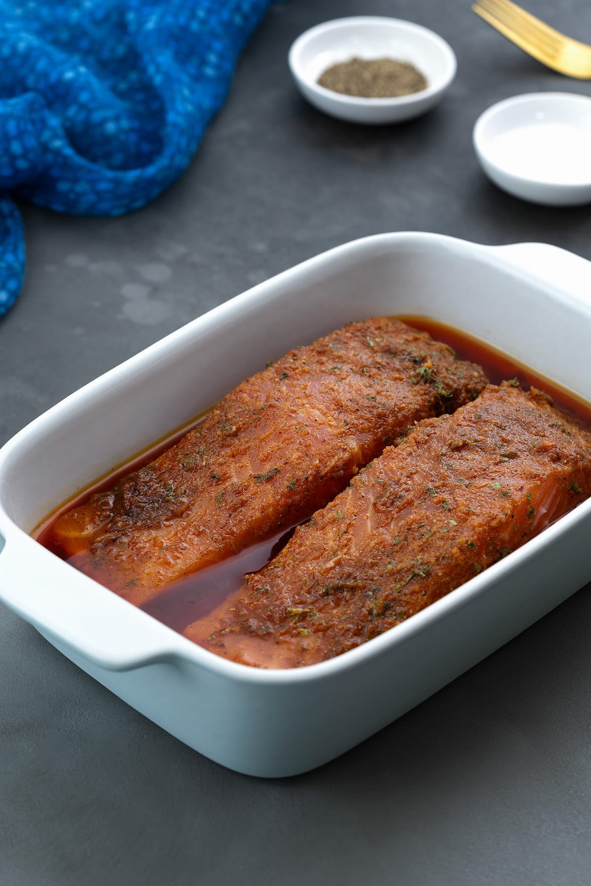 White baking tray filled with salmon fillets marinated in a freshly prepared homemade salmon marinade, accompanied by a small cup of salt and pepper and a blue towel.