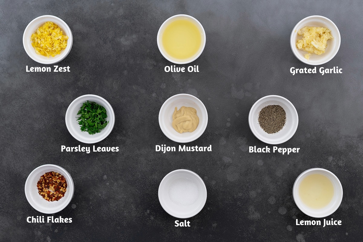 Shrimp marinade ingredients arranged on a gray table: lemon zest, olive oil, grated garlic, parsley leaves, Dijon mustard, black pepper, chili flakes, salt, and lemon juice.