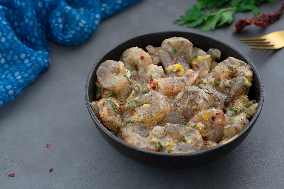 Marinated shrimp in a black bowl on a gray table, surrounded by a blue towel, a golden fork, parsley leaves, and red chilis.