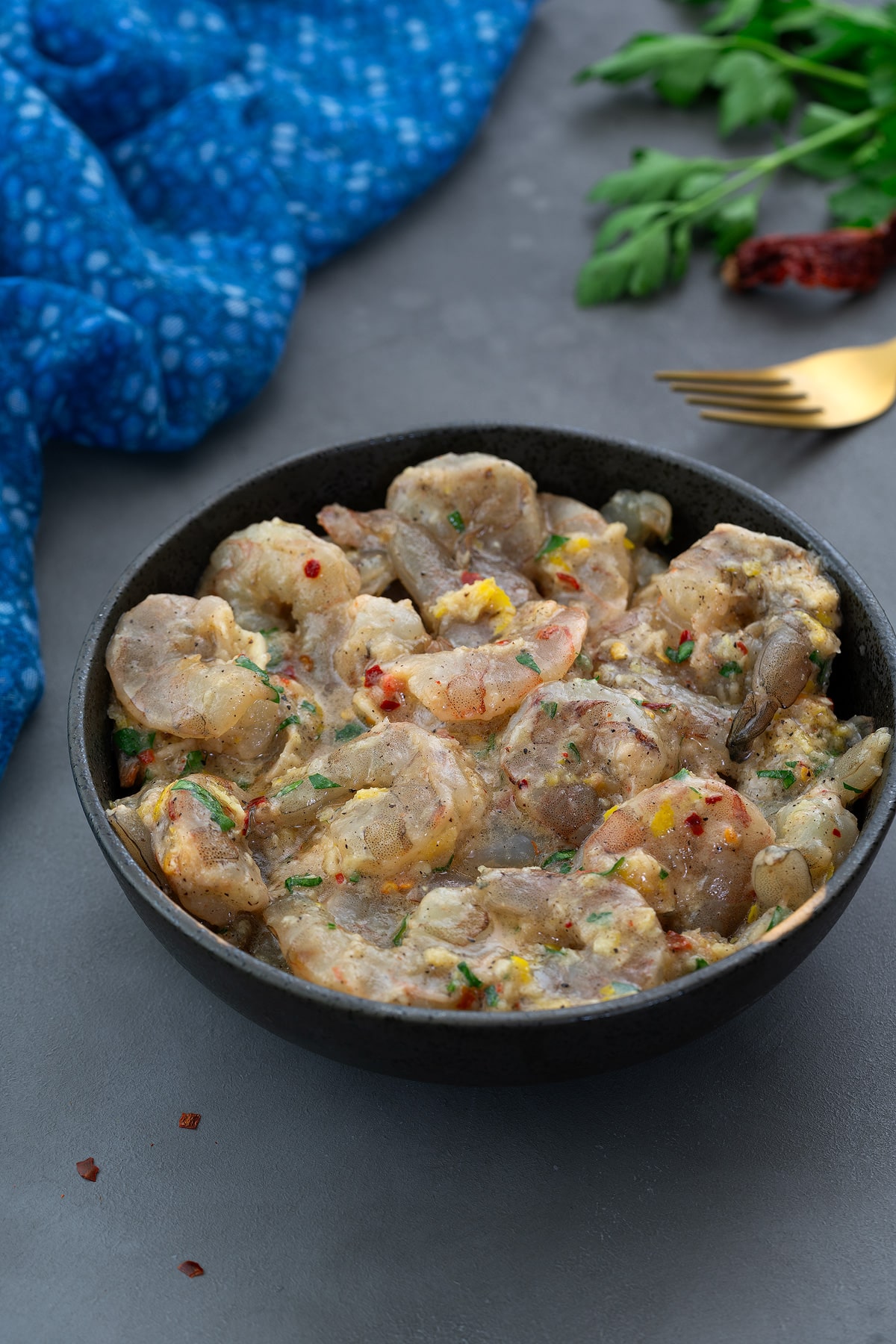 Marinated shrimp in a black bowl on a gray table, surrounded by a blue towel, a golden fork, parsley leaves, and red chilis.