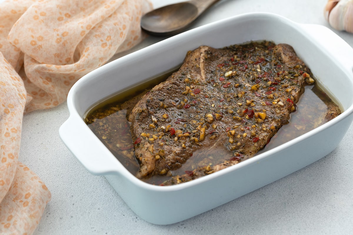 Steak marinade with steak cuts in a white dish on a white table with a towel, wooden spoon, and garlic.