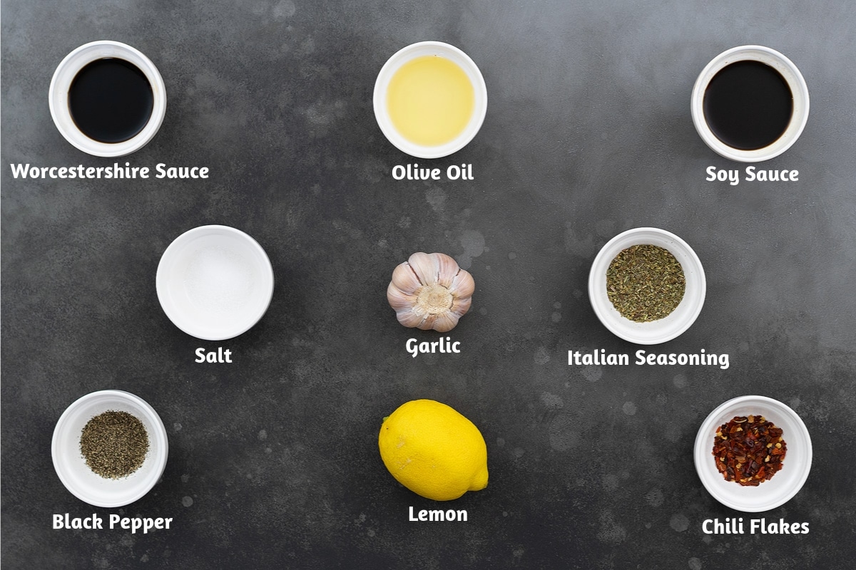 Ingredients for steak marinade recipe on a grey table: Worcestershire Sauce, Olive Oil, Soy Sauce, Salt, Garlic, Italian Seasoning, Black Pepper Powder, Lemon, Chili Flakes.