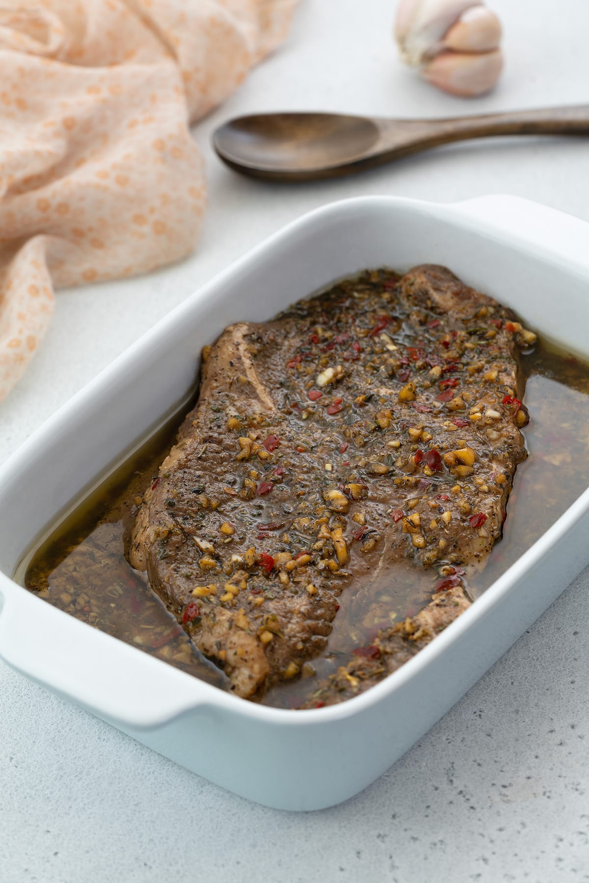 Steak marinade with steak cuts in a white dish on a white table with a towel, wooden spoon, and garlic.